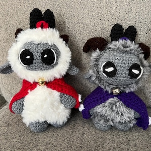 May include: Two crocheted plush figures, one white and gray with a red cape and the other gray with a purple cape. Both have large black eyes, small bells, and black horns.
