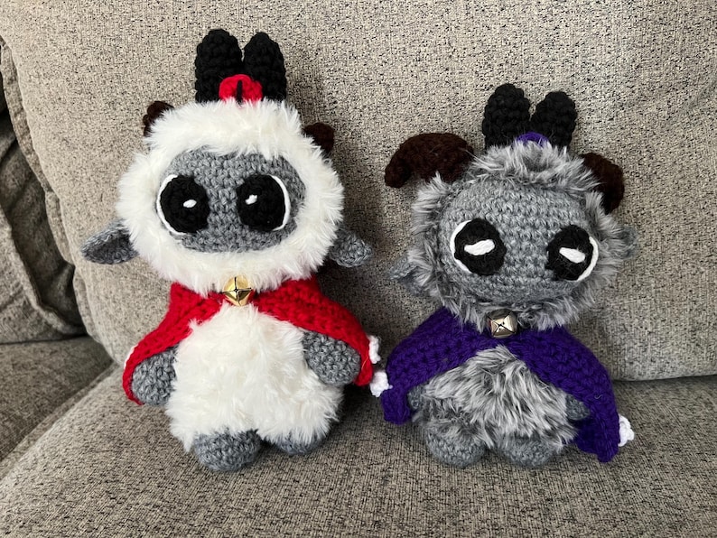 May include: Two crocheted plush figures, one white and gray with a red cape and the other gray with a purple cape. Both have large black eyes, small bells, and black horns.
