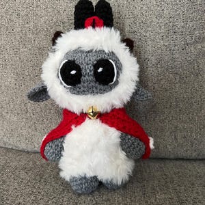May include: A handmade crochet lamb doll with a red cape and a gold bell. The lamb has a gray face and body, white fluffy fur around the face, and black horns. The doll is sitting on a gray surface.