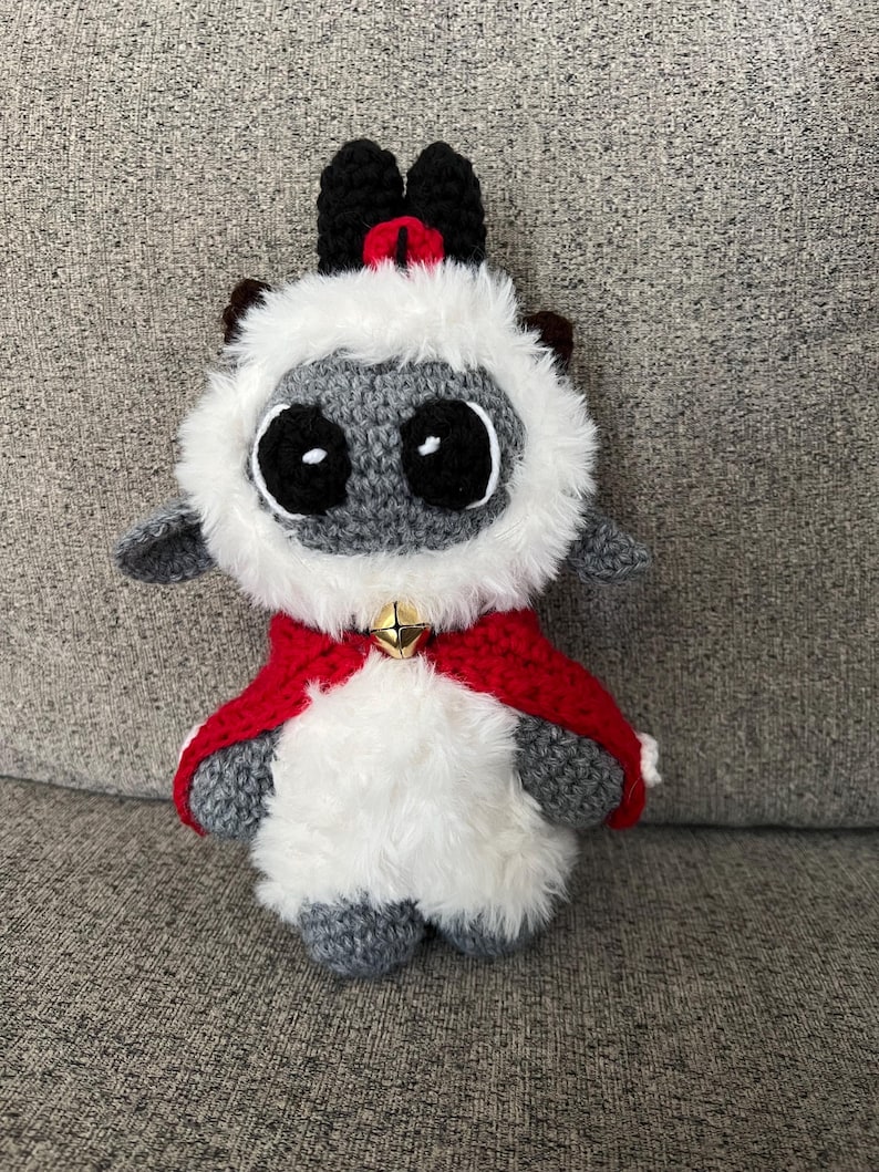 May include: A handmade crochet lamb doll with a red cape and a gold bell. The lamb has a gray face and body, white fluffy fur around the face, and black horns. The doll is sitting on a gray surface.