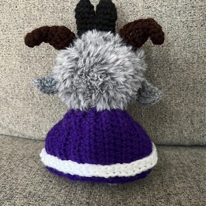 May include: A handmade crochet goat doll with a fluffy gray body, brown horns, and black ears. The doll wears a purple skirt with a white trim. The goat is a whimsical, soft toy.