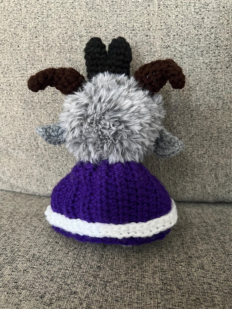 May include: A handmade crochet goat doll with a fluffy gray body, brown horns, and black ears. The doll wears a purple skirt with a white trim. The goat is a whimsical, soft toy.