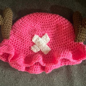 May include: A pink crocheted hat with brown crocheted antlers and a white crocheted X on the front.