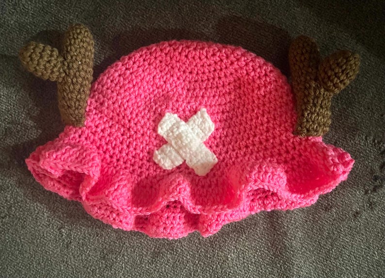 May include: A pink crocheted hat with brown crocheted antlers and a white crocheted X on the front.