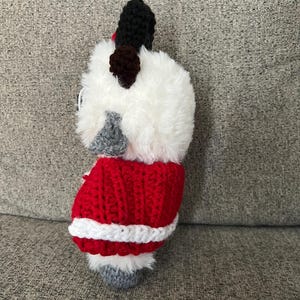 May include: A handmade, crocheted dog figurine. The dog has a fluffy white head, black ears, and a red and white crocheted coat. The dog's feet are gray. The figurine is a unique, handcrafted item.