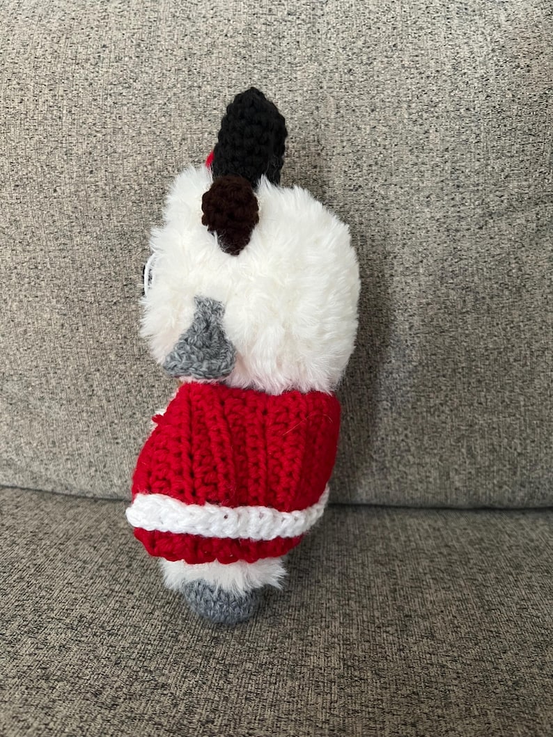 May include: A handmade, crocheted dog figurine. The dog has a fluffy white head, black ears, and a red and white crocheted coat. The dog's feet are gray. The figurine is a unique, handcrafted item.