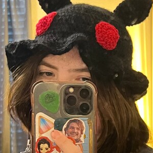 Puede incluir: Sombrero negro de ganchillo con detalles rojos. El sombrero tiene un dise&ntilde;o &uacute;nico con protuberancias en forma de orejas y un borde ondulado. Los detalles rojos son circulares.