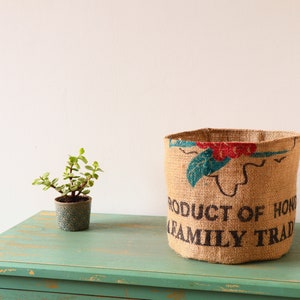Puede incluir: Un macetero de arpillera marrón con el texto "Producto de Honduras Tradición familiar" impreso en él. El macetero está sentado en una mesa de madera verde con una pequeña planta en maceta delante.