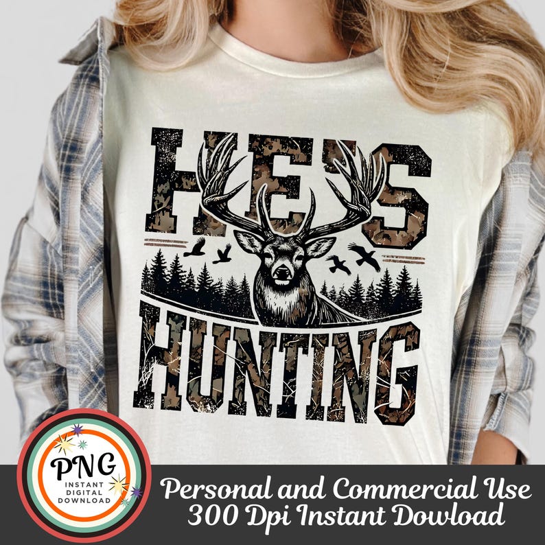 May include: White t-shirt with a camouflage pattern and the text "He's Hunting" in black. A black silhouette of a deer with antlers is in the center of the text.