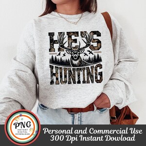 May include: A white sweatshirt with a camouflage print design featuring a deer with antlers and the text "He's Hunting".