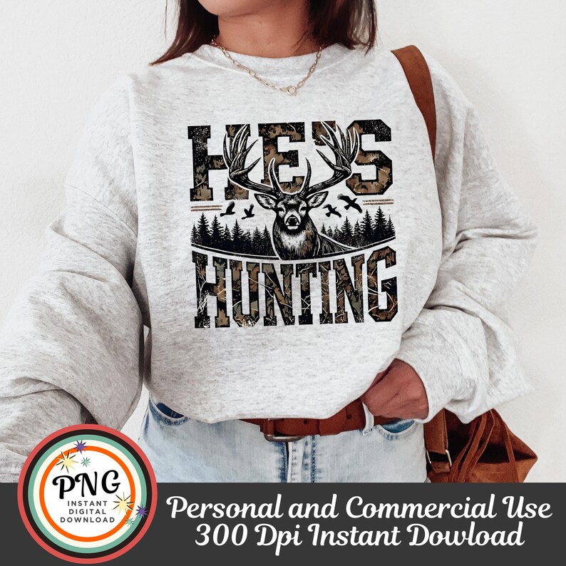 May include: A white sweatshirt with a camouflage print design featuring a deer with antlers and the text "He's Hunting".