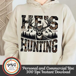 May include: A beige hoodie with a camouflage print design featuring a deer with antlers and the text "He's Hunting".