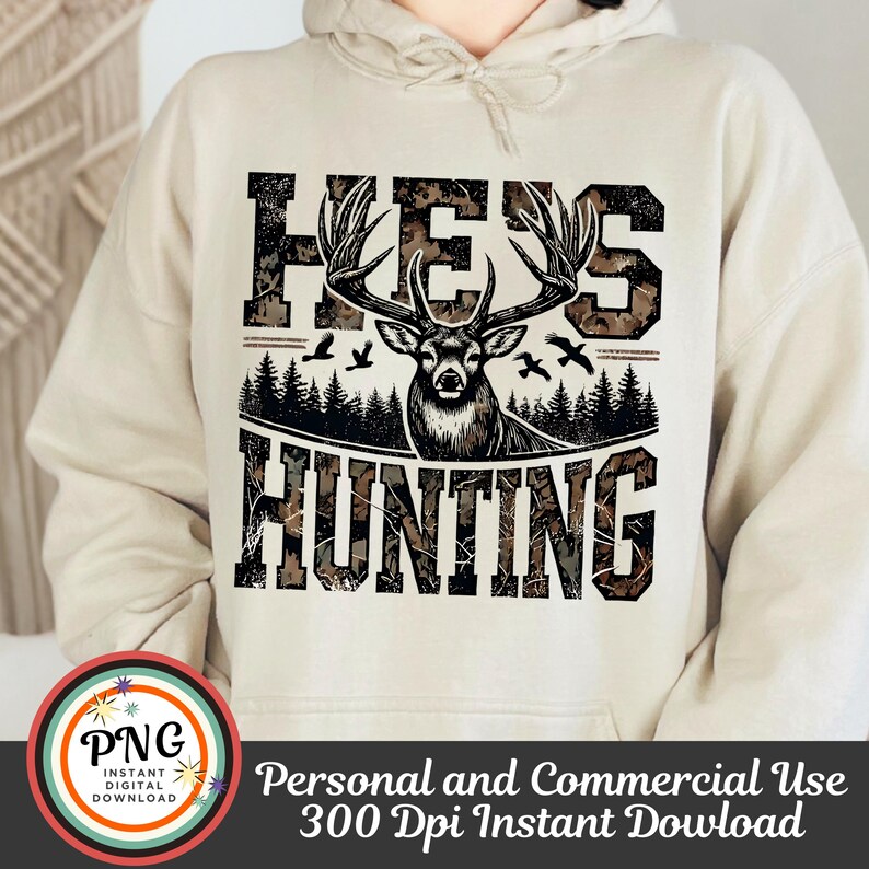 May include: A beige hoodie with a camouflage print design featuring a deer with antlers and the text "He's Hunting".