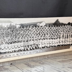 May include: A black and white photograph of a large group of people posing outdoors. The photo is mounted on a wooden frame with a metal ruler along the bottom. The group is in front of a building and trees.