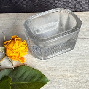 May include: A clear, faceted glass container with a rectangular shape. A dried yellow rose and green leaves are next to the container. The container is on a white wooden surface.