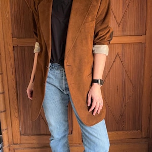 May include: A brown suede blazer with a classic notched lapel and two-button closure. The blazer is paired with a black V-neck shirt and light blue jeans. The sleeves are rolled up, revealing a cream-colored lining.