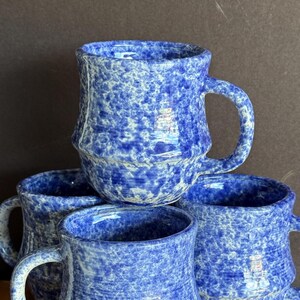 May include: Four blue ceramic mugs with a speckled pattern. The mugs have a cylindrical shape with a handle and a textured surface. The mugs are stacked on top of each other, showcasing their design.