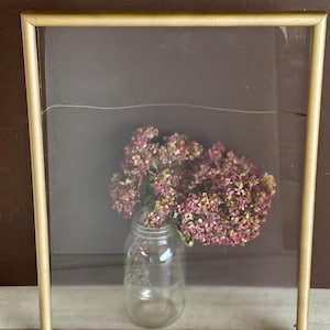 May include: A gold-framed picture with a clear glass pane. Inside, a glass jar holds a bouquet of dried, pink and beige flowers. The background is a dark brown color.