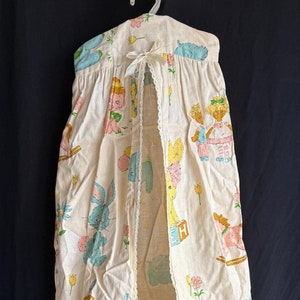 May include: A vintage baby nursery organizer made of cream-colored fabric, featuring a repeating pattern of pastel-colored bunnies, teddy bears, and children. The organizer has a lace trim and a white ribbon bow.