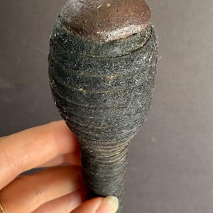 May include: A dark, textured object with a rounded top and a cylindrical body. The top is a darker, metallic color, while the body appears to be wrapped in a dark, fibrous material. The object is held by a hand.