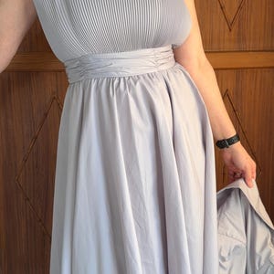 May include: A light gray dress with a pleated bodice and spaghetti straps. The dress has a cinched waist and a flowing skirt. A matching light gray jacket is held in the hand. The dress is a midi length.