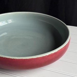 May include: A large, round ceramic bowl with a light blue interior and a deep red exterior. The bowl is sitting on a white surface.