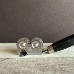 May include: A vintage knife sharpener with a black wooden handle and a silver-colored metal sharpening mechanism. The sharpener is marked "HAYES TOOL U.S.A.". The handle is angled, and the sharpener is resting on a wooden surface.
