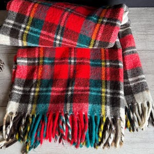 May include: A folded red, black, white, and teal plaid scarf with fringe. A pinecone and a sprig of evergreen are next to the scarf on a light gray wooden surface. The scarf is a winter accessory.