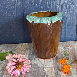May include: A handmade ceramic cup with a deep brown, textured body and a wavy, light turquoise glazed rim. The vessel features vertical indentations along its sides, creating a rustic, organic shape with a glossy finish on the exterior surface.