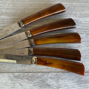 May include: Set of five steak knives with serrated blades and brown handles. The handles have a marbled appearance, transitioning from light to dark brown. Each knife has a silver bolster and the text "Robt. Hudson" is visible on the blade.