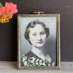May include: Vintage framed portrait of a woman with short, wavy hair. The portrait is in a rectangular gold-toned frame with ornate detailing. The woman is wearing a patterned dress. A pink and yellow flower is in the upper left corner.