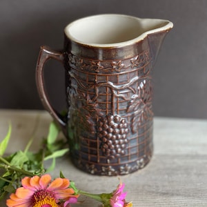 May include: A brown ceramic pitcher with a white interior and a grapevine design. The pitcher has a handle and a wide mouth. The pitcher is sitting on a wooden surface with pink flowers.