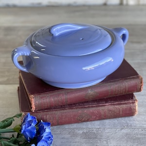 May include: A vintage, light blue ceramic casserole dish with a lid and two handles. The dish is sitting on top of three old, red books.