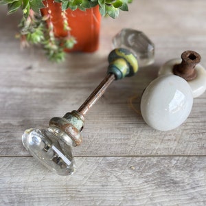 May include: Two antique glass doorknobs with metal stems. One knob is clear glass with a green and yellow swirl pattern. The other knob is clear glass. The knobs are on a light wood surface.