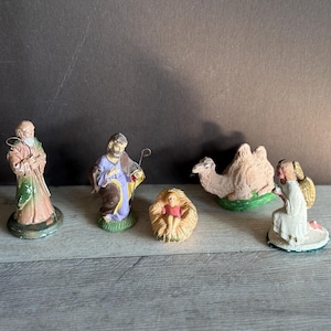 May include: A nativity scene set with five figurines: a shepherd, a man in a purple robe, a baby in a manger, a camel, and an angel. The figurines are made of painted plaster and are in a vintage style.