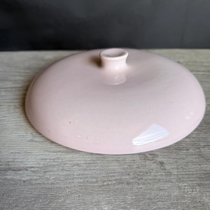 May include: A light pink ceramic lid with a small, rounded handle. The lid is circular and slightly domed, with a smooth, glossy finish. The lid is likely for a pot or other kitchenware.