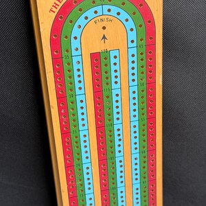 May include: Wooden cribbage board with a rectangular shape. The board features a track with red, green, and blue lanes, marked with numbers and holes for pegs. The text "THE U.S. PLAYING CARD CO." is printed on the board.