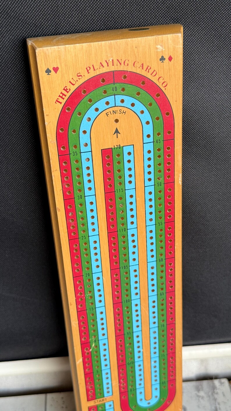 May include: Wooden cribbage board with a rectangular shape. The board features a track with red, green, and blue lanes, marked with numbers and holes for pegs. The text "THE U.S. PLAYING CARD CO." is printed on the board.