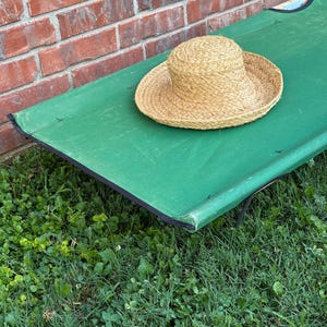 May include: A tan woven straw hat with a rounded crown and a wide brim resting on a rectangular green fabric cot with black trim. The cot is positioned on a patch of green grass next to a red brick wall, showcasing a simple outdoor setup.