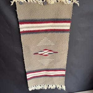May include: A woven wall hanging in shades of tan, red, white, and gray. The rectangular piece features horizontal stripes and a diamond pattern. The top edge has fringe and two dark gray buttons. The overall design has a Southwestern or Native American aesthetic.