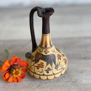 May include: A ceramic vase with a dark brown handle and neck, featuring a beige body adorned with black and brown illustrations of a deer and floral patterns. An orange flower with a yellow center rests beside the vase.