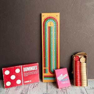 May include: A collection of vintage board games, including a wooden cribbage board with red, green, and blue tracks, a red dominoes box, a pink deck of Pinochle cards, and a box of red and tan game pieces.