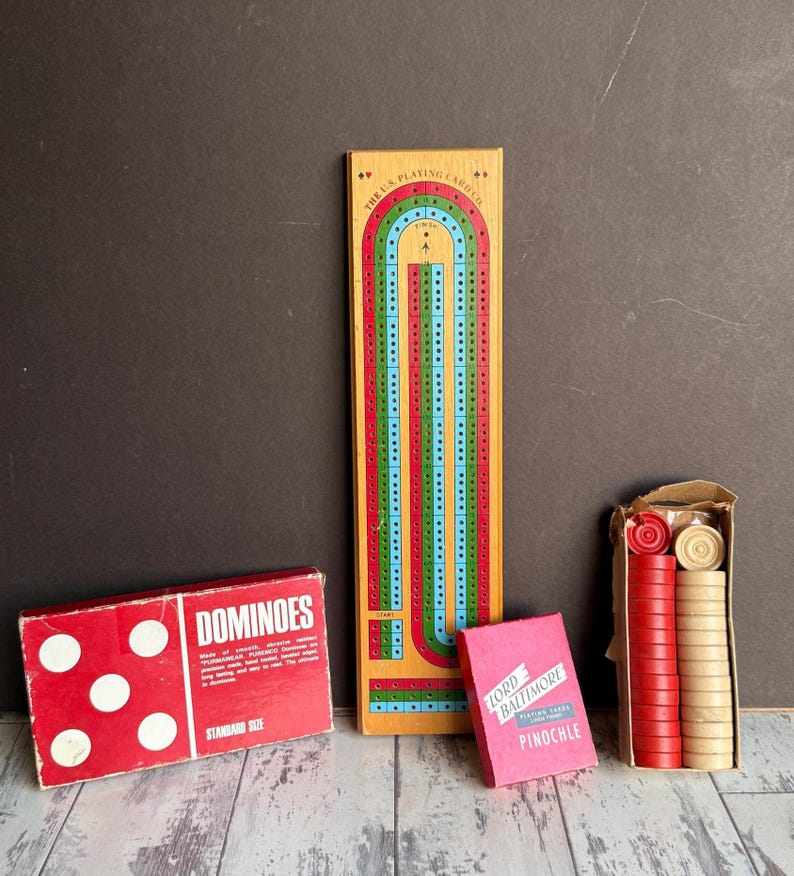 May include: A collection of vintage board games, including a wooden cribbage board with red, green, and blue tracks, a red dominoes box, a pink deck of Pinochle cards, and a box of red and tan game pieces.