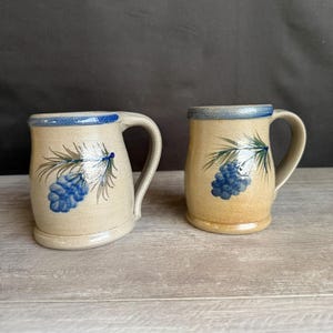 May include: Two ceramic mugs with a beige base and blue accents. Each mug features a blue and green botanical design. The rims and handles are blue. The mugs are displayed on a light wood surface.