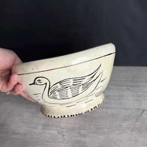 May include: Cream-colored ceramic bowl with a black swan illustration. The bowl has a wide, curved rim and a footed base with a decorative black border. The swan is depicted with detailed lines, adding a vintage aesthetic.
