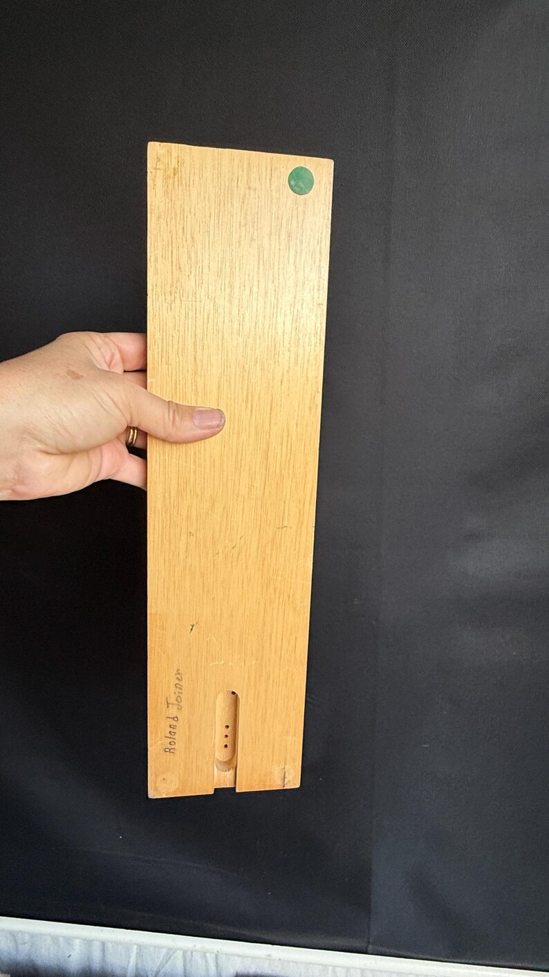 May include: A rectangular wooden board with a light brown finish, featuring a green circular sticker and a carved section. The board is held by a hand against a black background. The text "Roland Joiner" is visible.