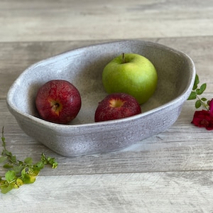 May include: An oval-shaped ceramic bowl with a speckled light gray finish. Inside the bowl are two red apples and one green apple. The bowl has a slightly irregular, hand-formed rim and a smooth, matte-textured surface throughout its exterior.