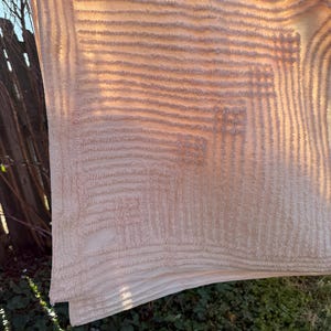 May include: Beige textured fabric with a geometric pattern, hanging outdoors. The fabric has a layered appearance with horizontal and square designs. The sunlight casts shadows, highlighting the texture and details of the material.