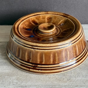 May include: A brown ceramic serving dish with a lid. The dish has a round shape with a ridged exterior and a small handle on the lid. The color is a mix of light and dark brown with some blue accents.