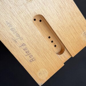 May include: Close-up of a light-colored wooden object with a rectangular shape and a carved-out section. The text "Roland Jerman" is handwritten in black ink on the wood. The object has several small holes.